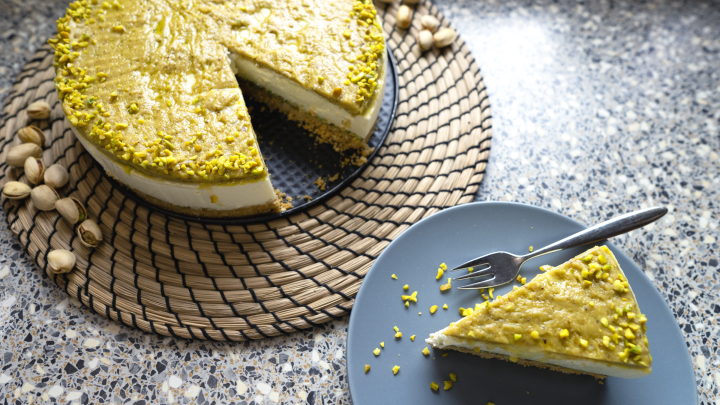 Ein cremiger Pistazien-Cheesecake mit einer leicht angerissenen Oberfl&auml;che liegt auf einem runden Untersetzer, umgeben von N&uuml;ssen.
