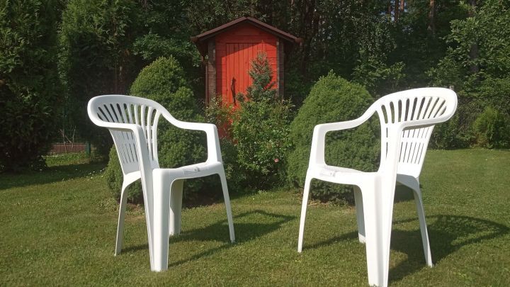 Zwei wei&szlig;e Kunststoffgartenst&uuml;hle stehen auf dem Rasen, umgeben von Str&auml;uchern und vor einem roten Gartenhaus.