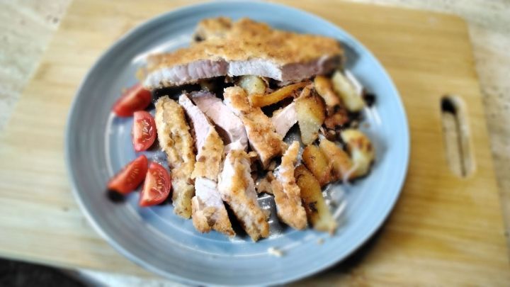 Auf einem blauen Teller liegen panierte Koteletts und knusprige Bratkartoffeln, umgeben von frischen Tomaten.