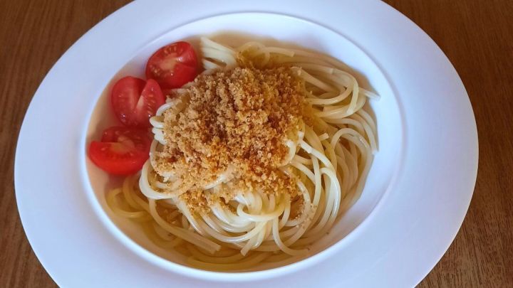 Spaghetti mit goldbraunen Semmelbr&ouml;seln und geschnittenen Tomaten auf wei&szlig;em Teller, pr&auml;sentiert auf holzfarbener Oberfl&auml;che.
