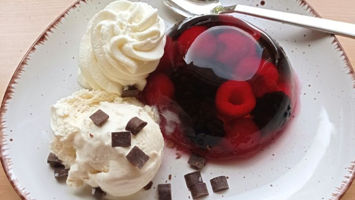 Eine Sch&uuml;ssel enth&auml;lt ein rotes Beerengelee, das mit Himbeeren gef&uuml;llt ist, daneben liegen zwei Kugeln Vanilleeis mit Schokoladensplittern.