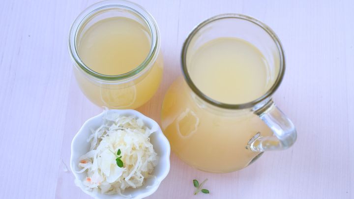 Ein Glas Sauerkrautsaft steht neben einer Schale mit frischem Sauerkraut auf einem hellen Tisch.