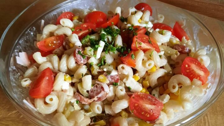 Ein Nudelsalat mit Tomaten, Mais und Kr&auml;utern ruht in einer Glassch&uuml;ssel auf einem Holztisch; ein Holzl&ouml;ffel liegt daneben.