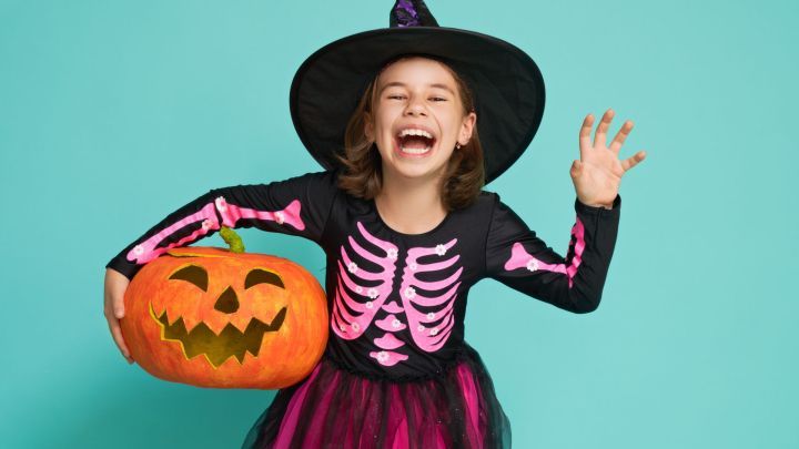 Ein Kind in Hexenkost&uuml;m lacht und h&auml;lt einen geschnitzten Halloween-K&uuml;rbis vor einem hellblauen Hintergrund.