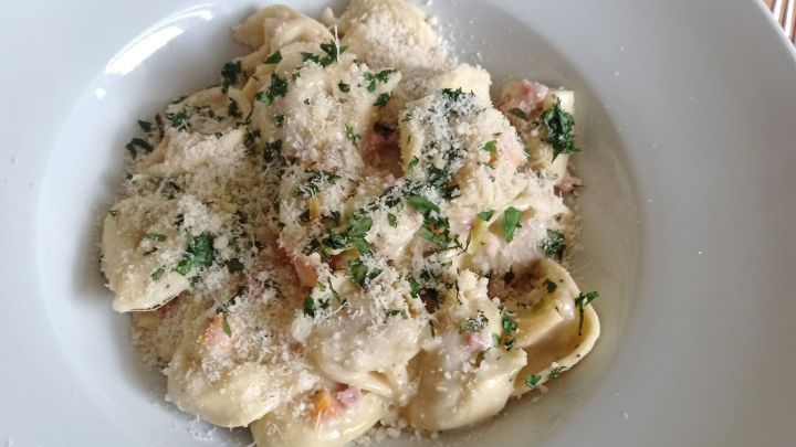 Tortelloni liegen in einer cremigen Speck-Sahne-So&szlig;e, bestreut mit K&auml;se und Kr&auml;utern, in einem wei&szlig;en Teller auf Holztisch.