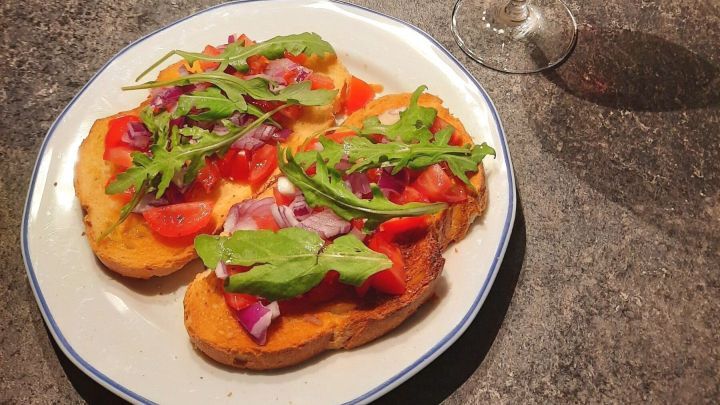 Bruschetta mit Tomaten, Rucola und Zwiebeln liegt auf einem wei&szlig;en Teller. Ein Glas Rotwein steht daneben auf einem dunklen Tisch.