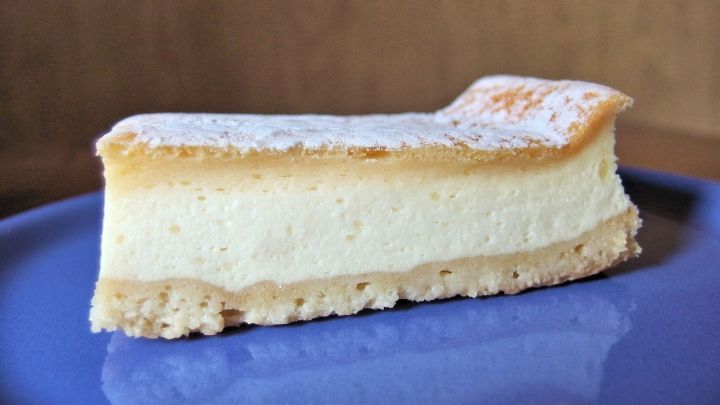 Ein K&auml;sekuchenst&uuml;ck liegt auf einem blauen Teller, leicht best&auml;ubt mit Puderzucker, locker und cremig in der Textur.