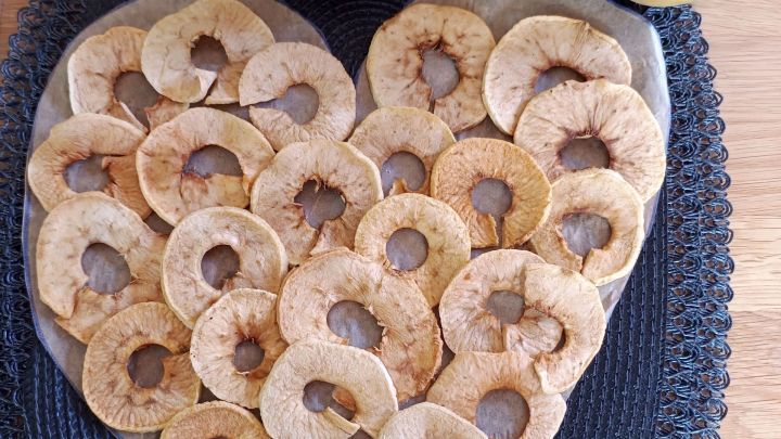 Getrocknete Apfelringe liegen in Herzform auf einem schwarzen Tischset, dar&uuml;ber sind drei frische &Auml;pfel arrangiert.