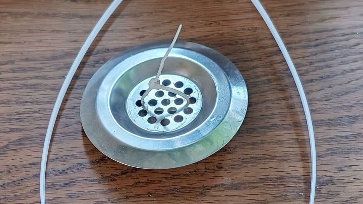 Ein metallisches Abflusssieb liegt auf einem Holztisch, fixiert mit einem Kabelbinder. Ein Zahnstocher steckt in den L&ouml;chern. Text: 17. Sept. 2024, 13:28.