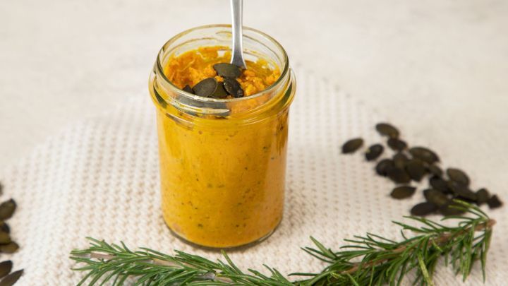 Ein Glas mit K&uuml;rbispesto, L&ouml;ffel steckt darin, steht auf beigefarbenem Tuch. Umgeben von K&uuml;rbiskernen und Rosmarinzweigen.