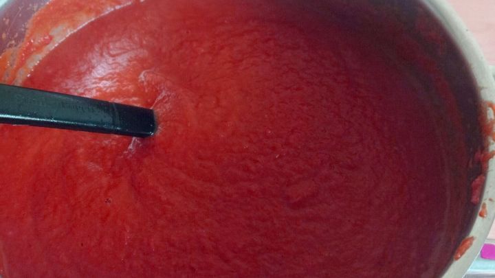 Topf voll roter Tomatenso&szlig;e; L&ouml;ffel r&uuml;hrt darin, auf Holzunterlage in K&uuml;chenumgebung.