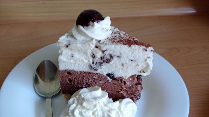 Ein St&uuml;ck Paradiescreme-Schnitte mit Kirsche obendrauf, serviert auf einem Teller mit Sahne und L&ouml;ffel, auf Holztisch.