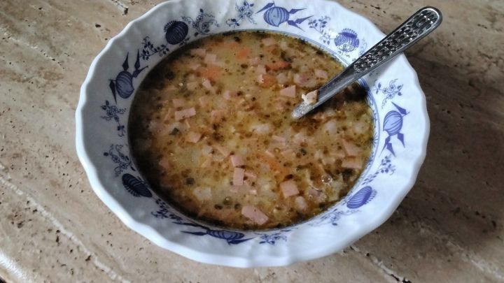 Eine keramikblaue Sch&uuml;ssel mit bunter Suppe, Kartoffelst&uuml;cken und Wurstw&uuml;rfeln steht auf einem Marmortisch, ein L&ouml;ffel darin.