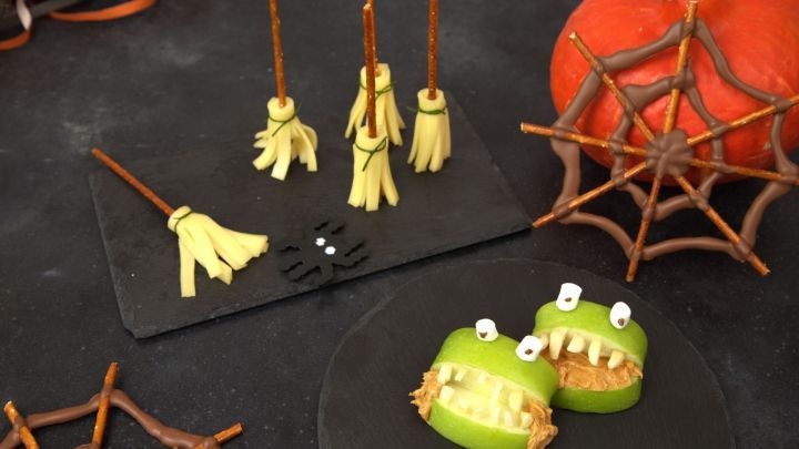 K&auml;sesticks als Besen, Apfelh&auml;lften mit Erdnussbutter als Monster neben einem K&uuml;rbis auf schwarzem Untergrund f&uuml;r Halloween-Snacks.