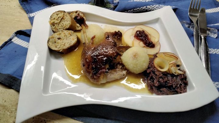 Gericht auf wei&szlig;em Teller: Entenhals gef&uuml;llt mit Kritharaki, dazu So&szlig;e, Kn&ouml;del, Apfelscheiben, Rotkohl; im Hintergrund Rotwein.