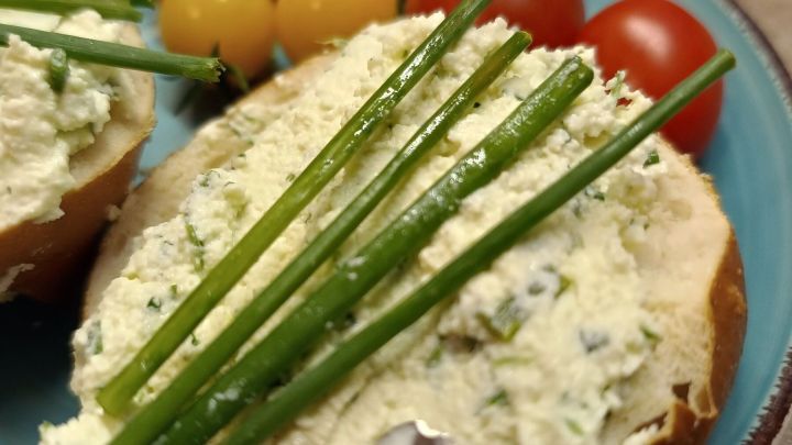 Br&ouml;tchenh&auml;lfte mit Kr&auml;uterfrischk&auml;se bestrichen, belegt mit Schnittlauch; im Hintergrund Tomaten auf blauem Teller. Datum: 26. Sept. 2024, 19:15.