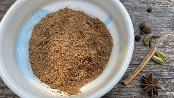 In einer Schale liegt Lebkuchengew&uuml;rz; daneben Zimtstange, Kardamom, Sternanis, angedeutet auf einem Holztisch.