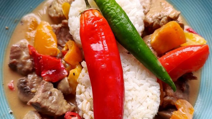 Fleischst&uuml;cke und Paprika in Erdnussso&szlig;e mit Reis, garniert mit roten und gr&uuml;nen Chilischoten auf einem blauen Teller.