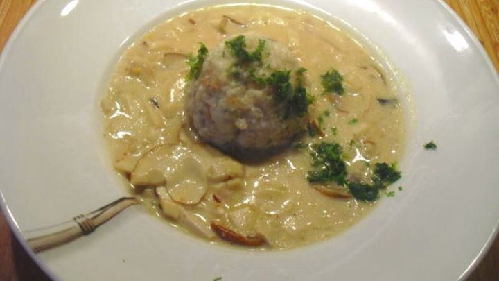 Kn&ouml;del ruht auf einem Teller in cremiger Steinpilzrahmso&szlig;e, garniert mit gr&uuml;ner Petersilie, auf einem Holzbrett pr&auml;sentiert.