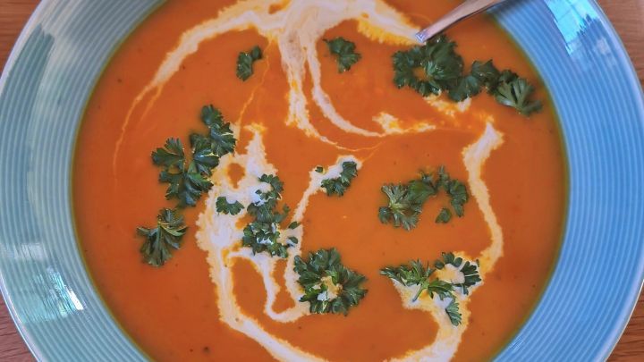 Eine Sch&uuml;ssel K&uuml;rbissuppe mit Sahneswirls und Petersilie auf einem Holztisch, umgeben von einem blauen Rand.
