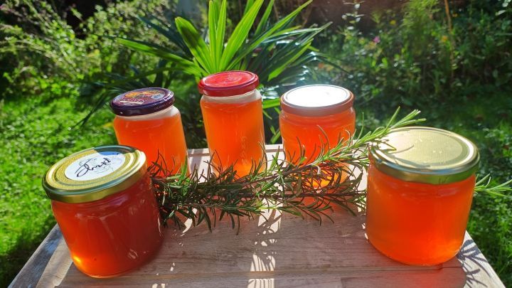 F&uuml;nf Gl&auml;ser Quittengelee stehen auf einem Holzbrett im Garten, dekoriert mit frischem Rosmarin. Ein Deckel zeigt das Wort &bdquo;Frucht&ldquo;.