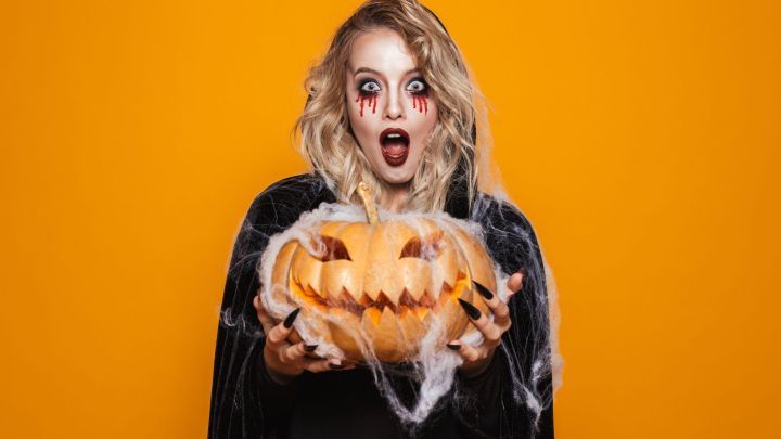 Eine Person in Halloween-Makeup h&auml;lt einen geschnitzten K&uuml;rbis voller k&uuml;nstlicher Spinnweben vor einem orangefarbenen Hintergrund.