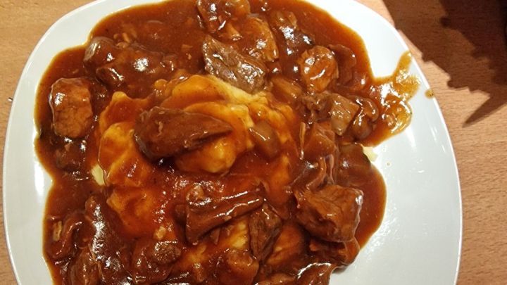 Ein Teller mit Gulasch in brauner So&szlig;e liegt auf einem Holztisch, dampfend und servierfertig, im warmen K&uuml;chenambiente.