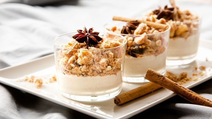 Gl&auml;ser mit Bratapfel-Crumble auf Zimtcreme stehen auf einem Tablett, garniert mit Zimtstangen und Sternanis, auf einem grauen Tuch.