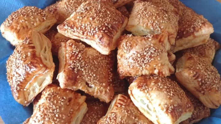 Bl&auml;tterteig-Kekse mit Sesam belegt liegen in einem blauen Tuch, arrangiert in einer Schale auf einem Holztisch.