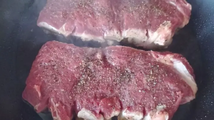 Zwei Rumpsteaks brutzeln in einer Pfanne, mit Pfeffer bestreut. Dampf umgibt das Fleisch in der K&uuml;che.