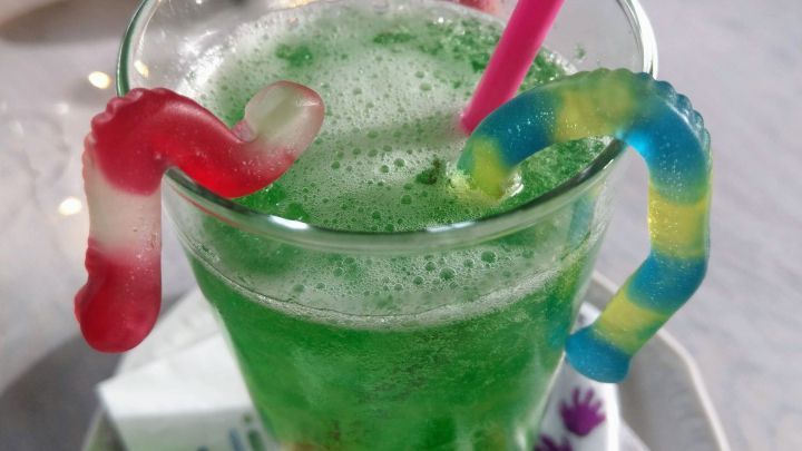 Ein gr&uuml;nes Getr&auml;nk mit Schaum und G&ouml;tterspeise, dekoriert mit bunten Gummiw&uuml;rmern, auf einem Teller mit Halloween-Serviette.