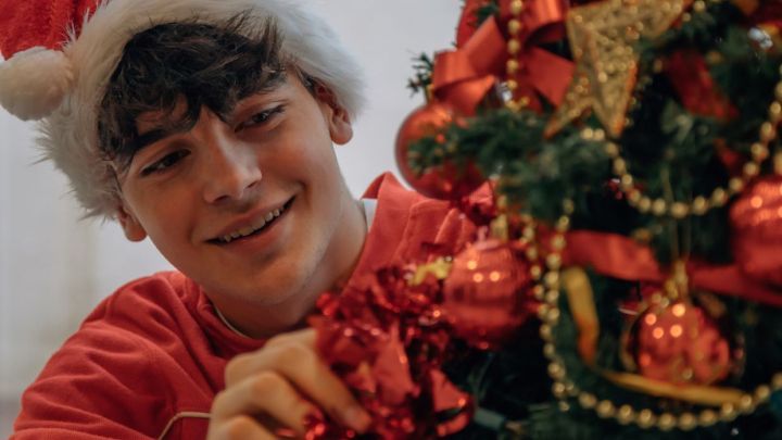 Ein Junge tr&auml;gt eine Weihnachtsm&uuml;tze und schm&uuml;ckt fr&ouml;hlich einen Weihnachtsbaum mit roten Kugeln und Schleifen im Raum.