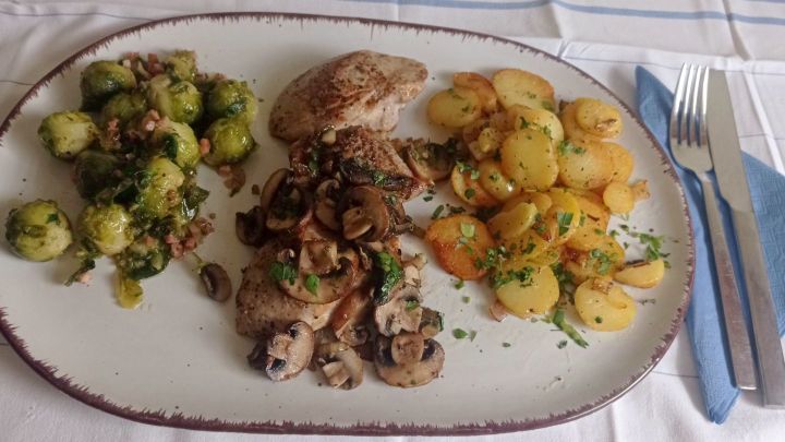 Auf einem Teller liegen Waidmanns-Filets mit Rosenkohl und Bratkartoffeln, getoppt mit frischen Kr&auml;utern und Pilzen auf den Filetscheiben.