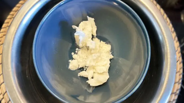 Wei&szlig;e Shea-Butterst&uuml;cke schmelzen in einer dunkelblauen Sch&uuml;ssel, die in einem Wasserbad steht, um Handcreme herzustellen.