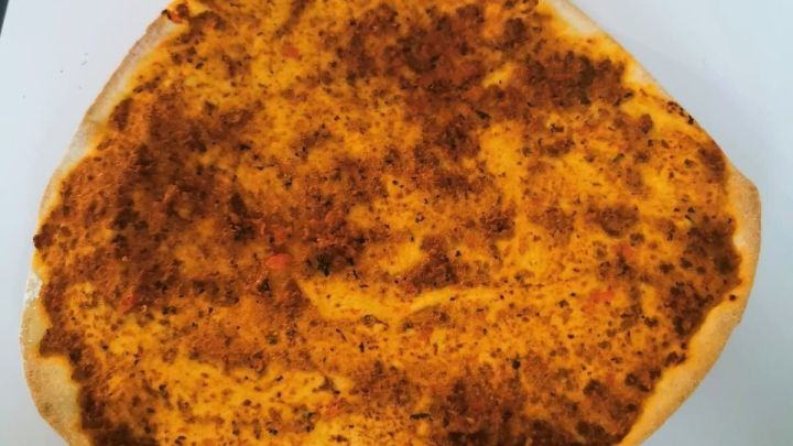 Eine gro&szlig;e, d&uuml;nne Lahmacun liegt auf einem wei&szlig;en Tisch und zeigt einen goldbraunen, gew&uuml;rzten Fleischbelag.