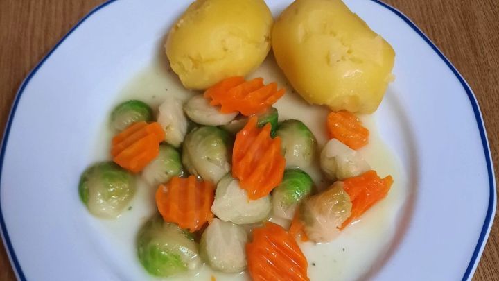 Rosenkohl liegt mit gekochten Kartoffeln und Karotten auf einem wei&szlig;en Teller, arrangiert auf einem Holztisch.