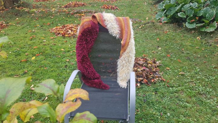 Eine gestrickte, herbstliche Stola liegt dekorativ auf einem Gartenstuhl, umgeben von buntem Herbstlaub auf gr&uuml;ner Wiese.