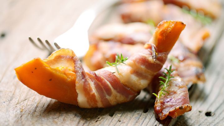 Ein K&uuml;rbisst&uuml;ck im Speckmantel ruht auf einem Holztisch, garniert mit frischem Thymian, w&auml;hrend eine Gabel danebenliegt.
