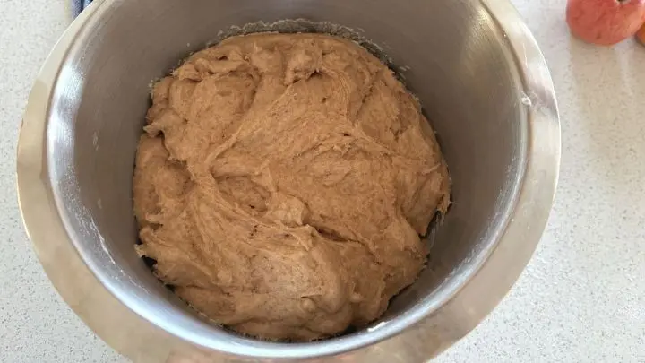 Ein Teig mit Zimt in einer Metallsch&uuml;ssel auf einer K&uuml;chenarbeitsplatte neben einem roten Apfel und einem Geschirrtuch.
