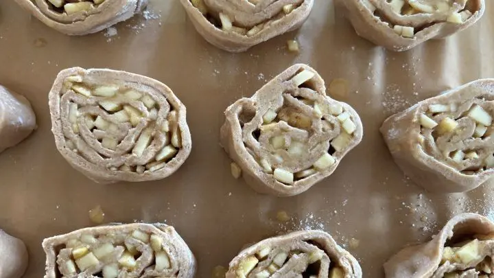 Schnecken aus Teig sind auf einem Backpapier ausgelegt. Sie zeigen Apfelst&uuml;cke und Zimt, bereit zum Backen.