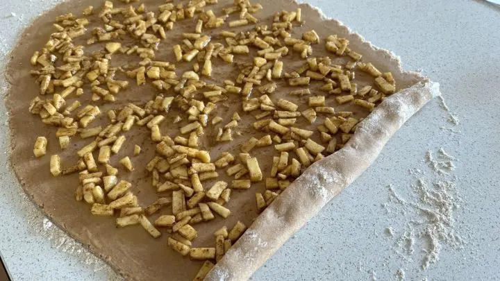 Ein ausgerollter Teig auf einer Arbeitsfl&auml;che wird zu Apfel-Zimt-Schnecken eingerollt, er ist mit Apfelst&uuml;cken belegt.
