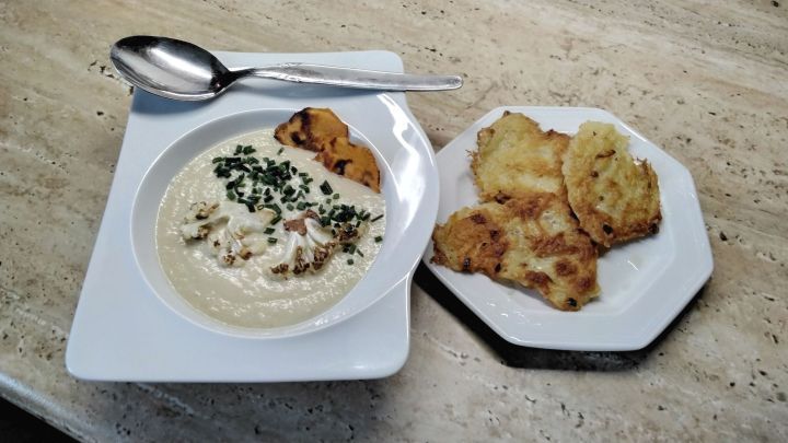 Eine Schale Blumenkohlcremesuppe mit Blumenkohlst&uuml;cken, S&uuml;&szlig;kartoffelscheiben und Schnittlauch, daneben Kartoffelpuffer auf einem Teller.
