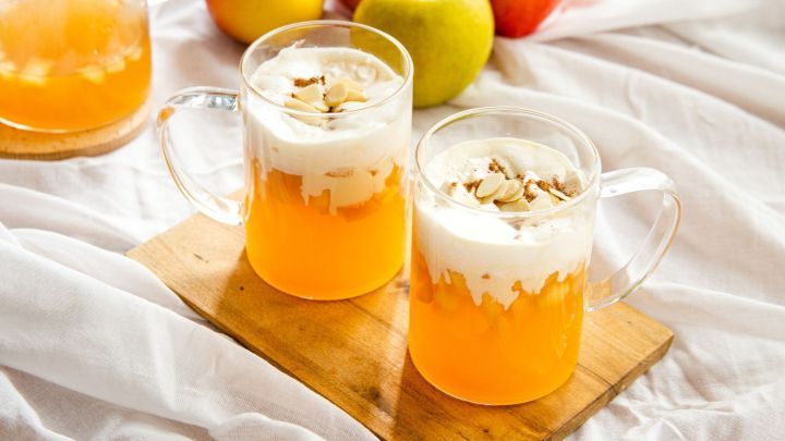 Zwei Glasbecher voll alkoholfreier Apfelpunsch mit Vanille-Sahne auf Holzbrett, dekoriert mit Mandeln, umgeben von &Auml;pfeln auf wei&szlig;em Tuch.