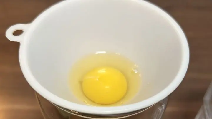 Schlage ein Ei an der Kante des Glases ohne Trichter auf. Kippe nun den Eiinhalt in den Trichter. Jetzt flie&szlig;t das Eiklar aus dem Trichter unten heraus. Das Eigelb ist aber ein bisschen zu dick und passt nicht durch. Perfekt! Denn nur das brauchen wir f&uuml;r unser Rezept.