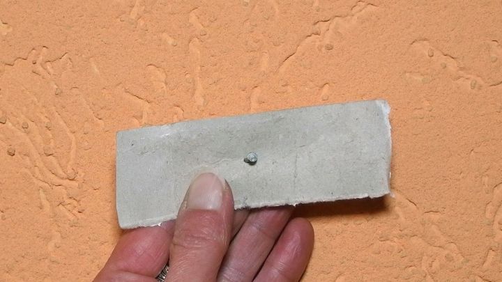 Eine Hand h&auml;lt einen schmalen, flachen Streifen, der an einer orangefarbenen Wand mit einem kleinen Nagel befestigt ist.