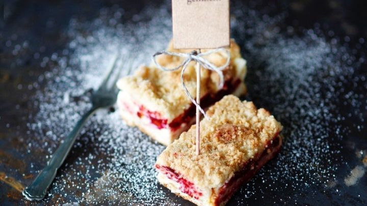 Ein Kirsch-Streuselkuchen mit Pudding liegt auf einem dunklen Teller, dekoriert mit Puderzucker und einem dekorativen Schild.