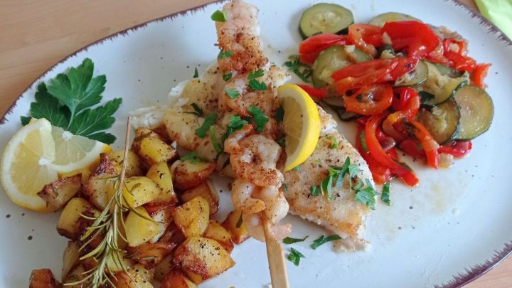 In der Pfanne gebratener Kabeljau mit Kartoffel- und Gem&uuml;sebeilage. Das Fischfilet wird noch mit dem kurz gebratenen Garnelen-Spie&szlig; belegt.