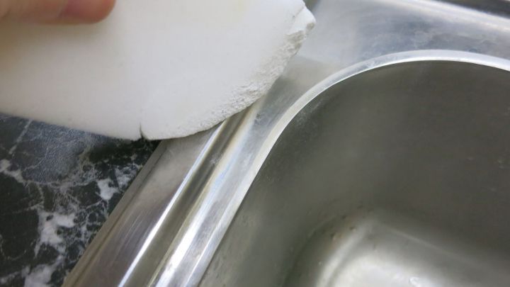 Eine Hand h&auml;lt einen Radierschwamm, der auf eine Edelstahlsp&uuml;le gerieben wird, um sie zu reinigen und wie neu erscheinen zu lassen.