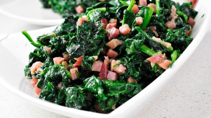 Rahmspinat wird in einer wei&szlig;en Sch&uuml;ssel pr&auml;sentiert, angereichert mit ger&auml;ucherten Schinkenw&uuml;rfeln und Zwiebeln.