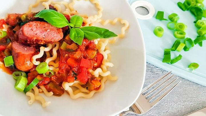 Ein Teller mit Wurstgulasch und Teigwaren steht auf einem Tisch, garniert mit frischen Kr&auml;utern und frischen Zwiebeln.